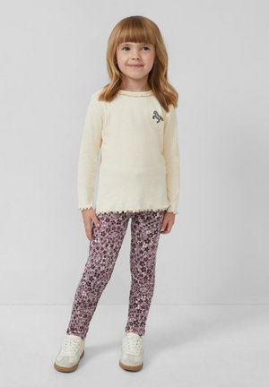 Kind dat een geribbelde crèmekleurige longsleeve top met een gerimpelde zoom en een paardenlogo draagt, in combinatie met paarse leggings met bloemenpatroon en witte sneakers.