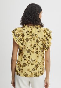Blusa gialla con fiori, maniche a volant, motivo floreale nero, tessuto testurizzato, dettaglio con apertura a chiave sul retro e vestibilità rilassata.