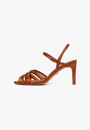 Sandalo marrone con cinturini, tacco alto, fibbia alla caviglia e punta aperta, texture liscia e tacco sottile affusolato su sfondo bianco.
