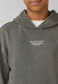 Persona llevando una sudadera con capucha gris con texto blanco que dice "EL ESPÍRITU DEL OESTE SALVAJE. El encanto del país y el oeste cautivará tu corazón."
