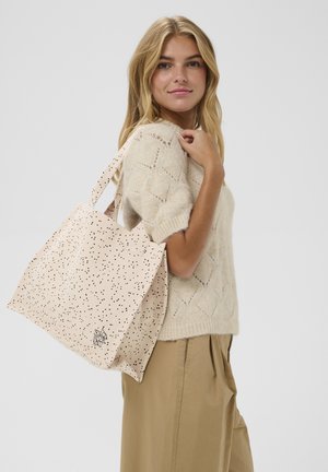 Szőke nő bézs színű kötött pulóverben és khaki nadrágban nagy bézs színű táskával, fekete foltmintával a vállán.