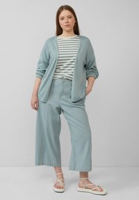 Hellblauer Cardigan, gestreiftes Hemd in gedämpften Tönen, lässige hellblaue Hose und sandfarbene Riemchensandalen. Weicher Stoff und entspannter Sitz.