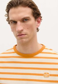 Camiseta a rayas en crema y naranja, con un cuello redondo acanalado y un pequeño parche con el logo en el lado izquierdo del pecho. Textura de tela suave.