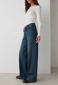 Wide-leg dark blue denim jeans with a high waist, featuring five pockets and a subtle faded pattern. Paired with a fitted white long-sleeve top.