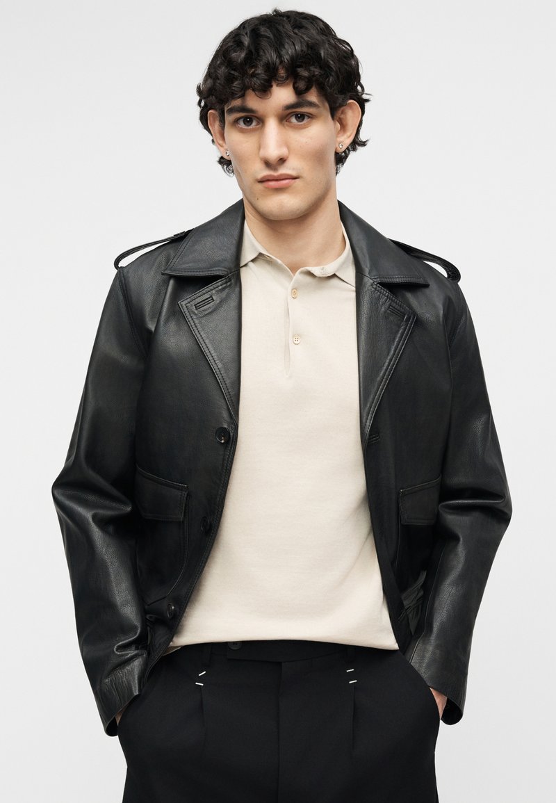 Young man with curly dark hair wears a black leather jacket over a beige polo shirt and black pants, standing with hands in pockets.