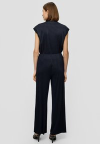 Navyblauwe, geribbelde jumpsuit met kapmouwen, elastische tailleband, ontwerp met brede pijpen; gecombineerd met zwarte hakken. Achteraanzicht weergegeven.
