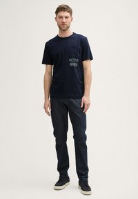 Camiseta de algodón azul marino con un bolsillo en el pecho y un diseño impreso en texto verde claro. Combinada con jeans de mezclilla oscura y zapatillas negras.