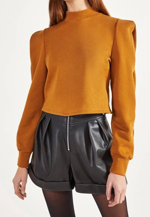 Femme portant un haut moutarde à manches longues avec épaules bouffantes et un short noir taille haute en simili cuir avec une fermeture éclair à l'avant.