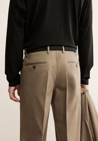 Pantaloni eleganti beige con vestibilità su misura, dotati di due tasche posteriori con chiusura a bottone, indossati con una camicia nera a maniche lunghe.