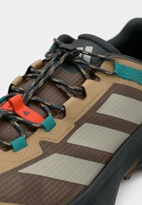 Chaussures de sport avec une tige marron texturée, des accents beige, des lacets noirs et bleu sarcelle, et des bandes blanches proéminentes sur le côté.