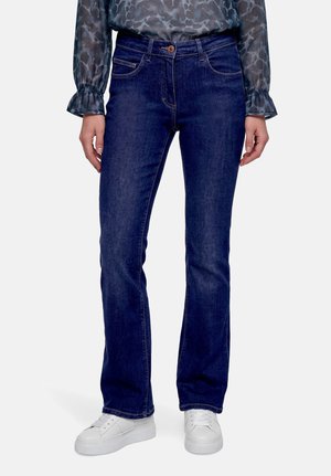 Femme portant un jean droit bleu foncé, des baskets blanches et un top transparent à manches longues avec un motif bleu-gris, debout devant un fond uni.