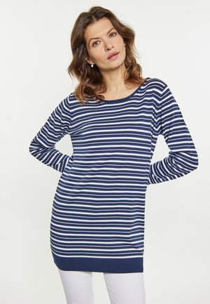 Kvinde iført en marineblå og hvidstribet bluse med lange ærmer og hvide bukser, stående med hænderne bag ryggen, neutral baggrund.