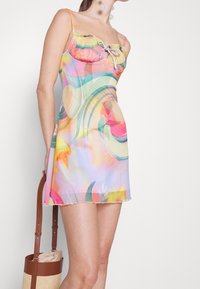 Vícebarevné mini šaty vyrobené z lehkého materiálu s volánkovým výstřihem, tenkými ramínky a abstraktními květinovými vzory. Detaily na třepení lemu.