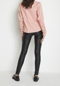 Blouse rose clair avec des manches longues froncées, associée à un legging en cuir synthétique noir et des chaussures à plateforme noires, vue de dos.