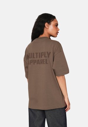 T-shirt in cotone marrone con maniche corte, con la scritta "MULTIPLY APPAREL" stampata in marrone scuro sul retro. Vestibilità rilassata e colletto a girocollo.