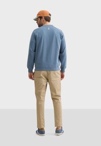 Sudadera azul claro con puños acanalados, pantalones beiges y zapatillas deportivas azules con detalles marrones. Llevado con una gorra naranja. Vista desde atrás.