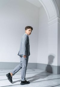 Szary garnitur z teksturowanej tkaniny, o dopasowanym kroju. Czarne buty na masywnej podeszwie. Stoi na marmurowej podłodze, ustawiony pod kątem do jasnej ściany.