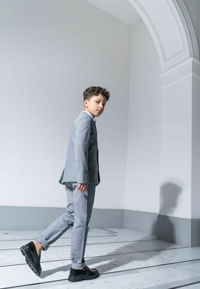 Szary garnitur z teksturowanej tkaniny, o dopasowanym kroju. Czarne buty na masywnej podeszwie. Stoi na marmurowej podłodze, ustawiony pod kątem do jasnej ściany.