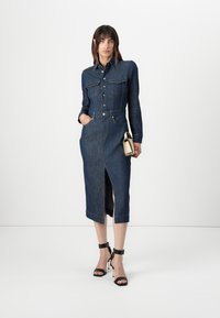 Mørk denim skjortekjole med innsvingt midje, lange ermer, trykknapper, frontlommer og en høy splitt til låret. Kombinert med svarte sandaler.