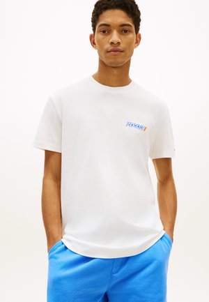 Bărbat tânăr purtând un tricou alb cu logo-ul Tommy și pantaloni albaștri, cu mâinile în buzunare, stând în fața unui fundal simplu și deschis la culoare.