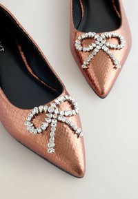 Ballerines pointues en rose gold avec une finition texturée et un ornement de nœud en rhinestones sur le devant. Doublure intérieure lisse noire.