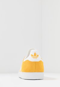 Gula mockaskor med vita accenter. Har en texturerad sula och en logga på hälen. Bakifrån visar de en ren design.