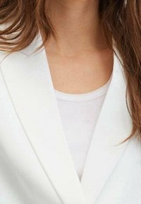 Kobieta w białym żakiecie nosi białą bluzkę z okrągłym dekoltem i prążkowaniem, z długimi brązowymi włosami częściowo widocznymi.