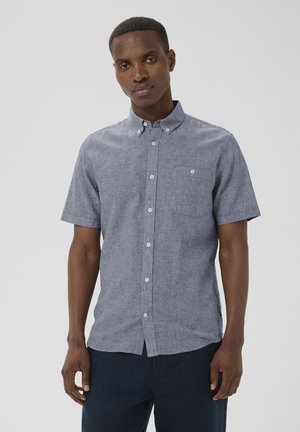 Mand iført lyseblå kortærmet skjorte med brystlomme, stående foran en ensfarvet grå baggrund.