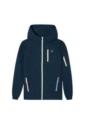Niño caminando con una chaqueta azul marino, jersey crema sobre una camisa azul, vaqueros azul claro y zapatillas oscuras contra un fondo liso.