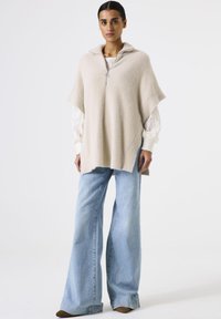 Woman wearing a beige zip-up poncho over a white textured blouse and light blue wide-leg jeans standing against a plain white background.