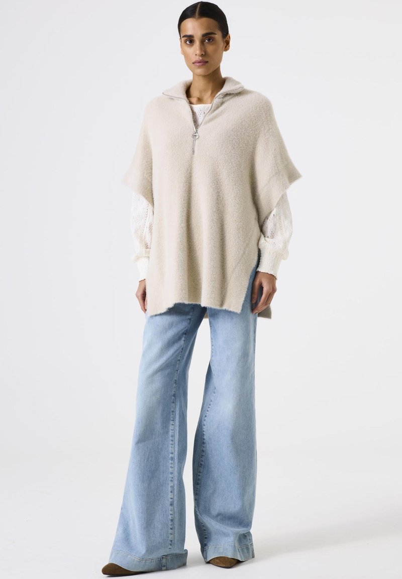 Woman wearing a beige zip-up poncho over a white textured blouse and light blue wide-leg jeans standing against a plain white background.