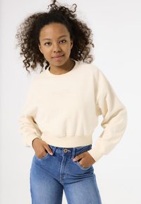 Sudadera corta de color crema en tejido suave, con puños y dobladillo acanalados. Combinada con vaqueros de mezclilla azules de cintura alta con cierre de botón.