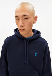 Marineblauwe hoodie van zachte stof, voorzien van een voorkantzak en trekkoorden, met een klein blauw logo op de linkerborst.