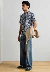 Μπλε ναυτικό T-shirt με λευκό λουλουδάτο σχέδιο, μπεζ πουλόβερ περασμένο στον ώμο, φαρδιά μπλε τζιν και μπλε ναυτικά αθλητικά παπούτσια με λευκές λεπτομέρειες.