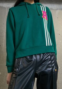 Felpa verde con strisce bianche di lato, con una lettera 'A' ricamata in rosa. Indossata con pantaloni in pelle nera lucida.