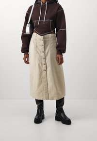 Ruskea lyhyt huppari, jossa valkoiset ompeleet, yhdistettynä beigeen nappihameeseen ja mustiin platformnilkkureihin. Silkkinen kangas, rento istuvuus.