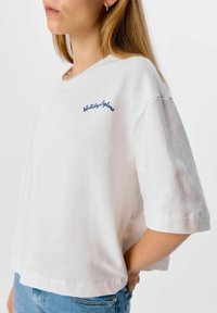 Camiseta de algodón blanca con un corte relaxed, mangas cortas, y bordado en azul marino "Explora las Vacaciones" en el lado izquierdo. Textura ligera.