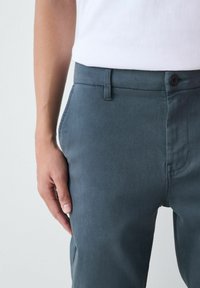 Közeli kép egy személyről, aki sötétszürke nadrágot és fehér inget visel, bal keze pedig lazán az oldalán pihen.