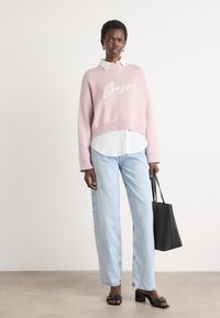 Rosa strikkegenser med "Boss" i hvit kursiv, laget over en hvit skjorte med krage. Lyseblå straight-leg jeans og svart håndveske, med svarte hælsandaler.