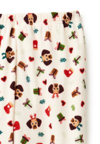 Fleece pyjamabroek in crème met een herhaald patroon van tekenfilmhondjes, cadeaus, harten en kerstmotieven in rood, groen en bruin.