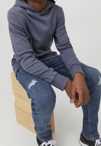 Sudadera gris con una superficie texturizada, combinada con jeans azules desgastados. El atuendo incluye zapatillas negras y blancas y se encuentra sobre un bloque de madera.