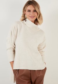 Cremfarbener Rollkragenpullover mit langen Ärmeln, weicher Textur und lockerer Passform. Kombiniert mit braunen Lederoptik-Hosen.