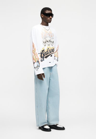 Άτομο που φοράει υπερμεγέθη λευκό φούτερ με γραφικά κρανίου και φλόγας, ανοιχτό μπλε τζιν με φαρδιά πόδια, μαύρα και άσπρα παπούτσια και μαύρα γυαλιά ηλίου.