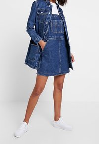 Vestido de mezclilla con un corpiño ajustado y falda, que presenta un bolsillo delantero y cierres de botones, combinado con una chaqueta de mezclilla larga. Zapatillas blancas.