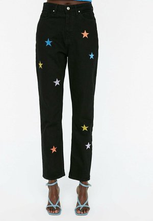 Schwarze Jeans mit geradem Bein und bunten bestickten Sternen, kombiniert mit hellblauen Riemchen-Sandaletten mit hohen Absätzen auf dunkler Haut.