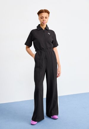 Combinaison de sport noire avec fermeture éclair à l'avant, manches courtes, taille élastique et bandes latérales blanches. Associée à des chaussures violettes sur un fond bleu.