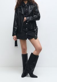 Persona che indossa una giacca di pelle nera, un vestito di jeans neri corto e stivali di pelle nera larghi su uno sfondo bianco semplice.