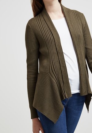 Femme portant un cardigan côtelé vert olive avec une chemise blanche et un jean bleu, vue du cou aux hanches sur un fond uni.