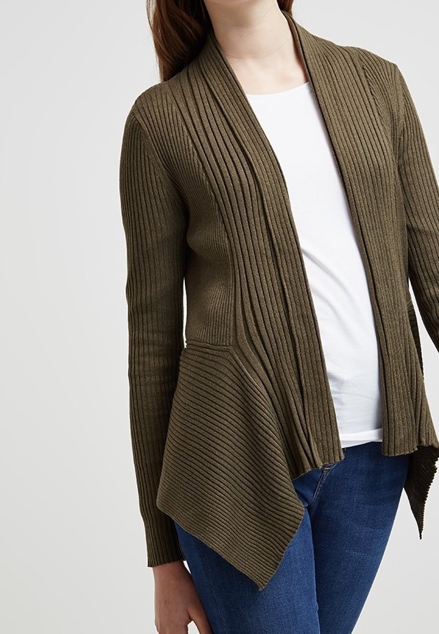 Femme portant un cardigan côtelé vert olive avec une chemise blanche et un jean bleu, vue du cou aux hanches sur un fond uni.