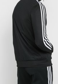 Mustapaita vetoketjulla, joka on valmistettu sileästä kankaasta ja jossa on kolme valkoista raitaa jokaisessa hihassa sekä ribbireunukset vyötäröllä.